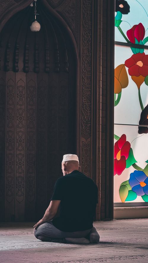 Free A man in traditional attire prays inside a mosque, near a colorful stained glass window. Stock Photo