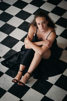A woman in a black dress sits on a black-and-white checkered floor, looking up thoughtfully.