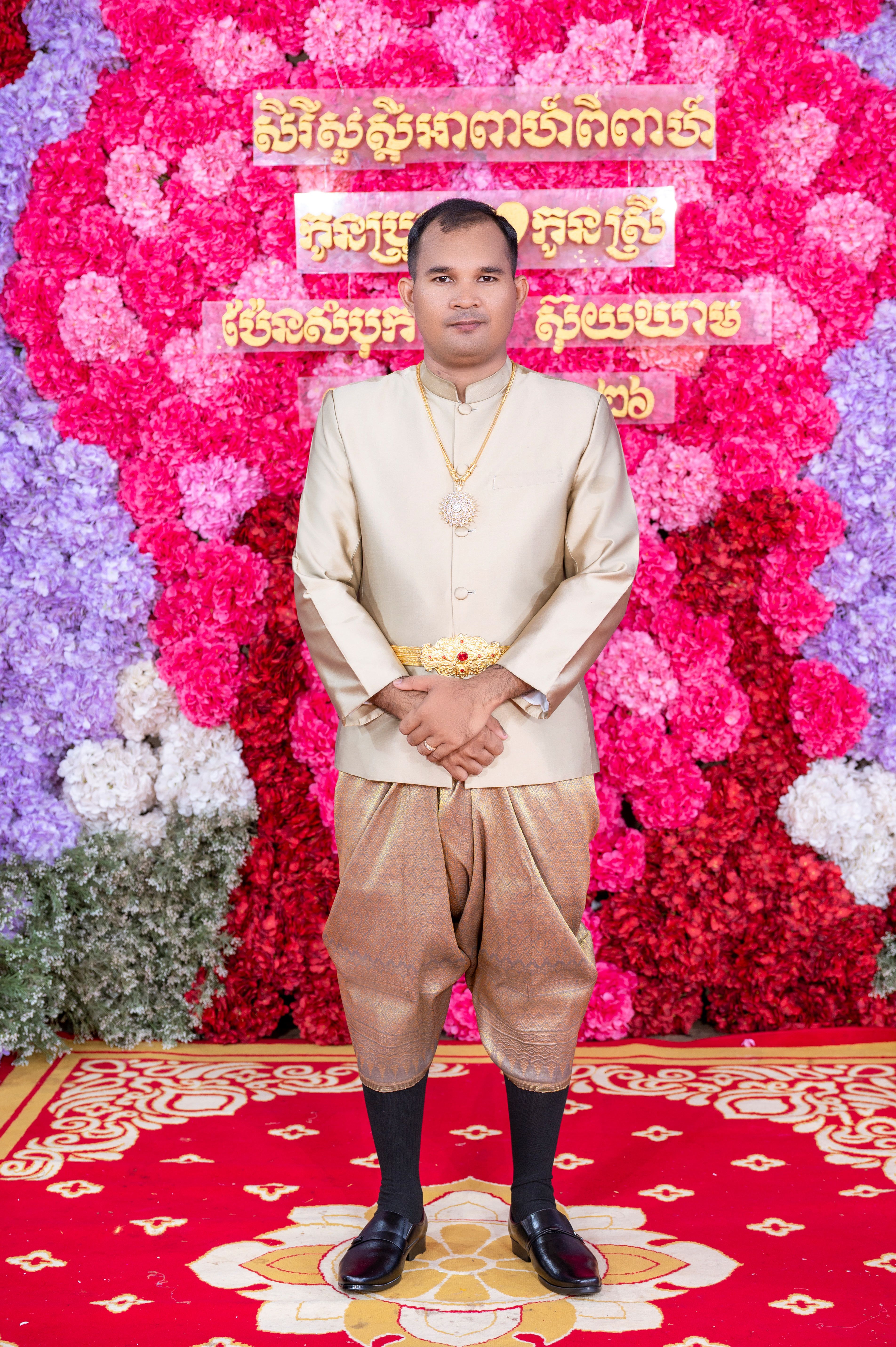 Man in elegant Cambodian wedding attire standing against a floral background.