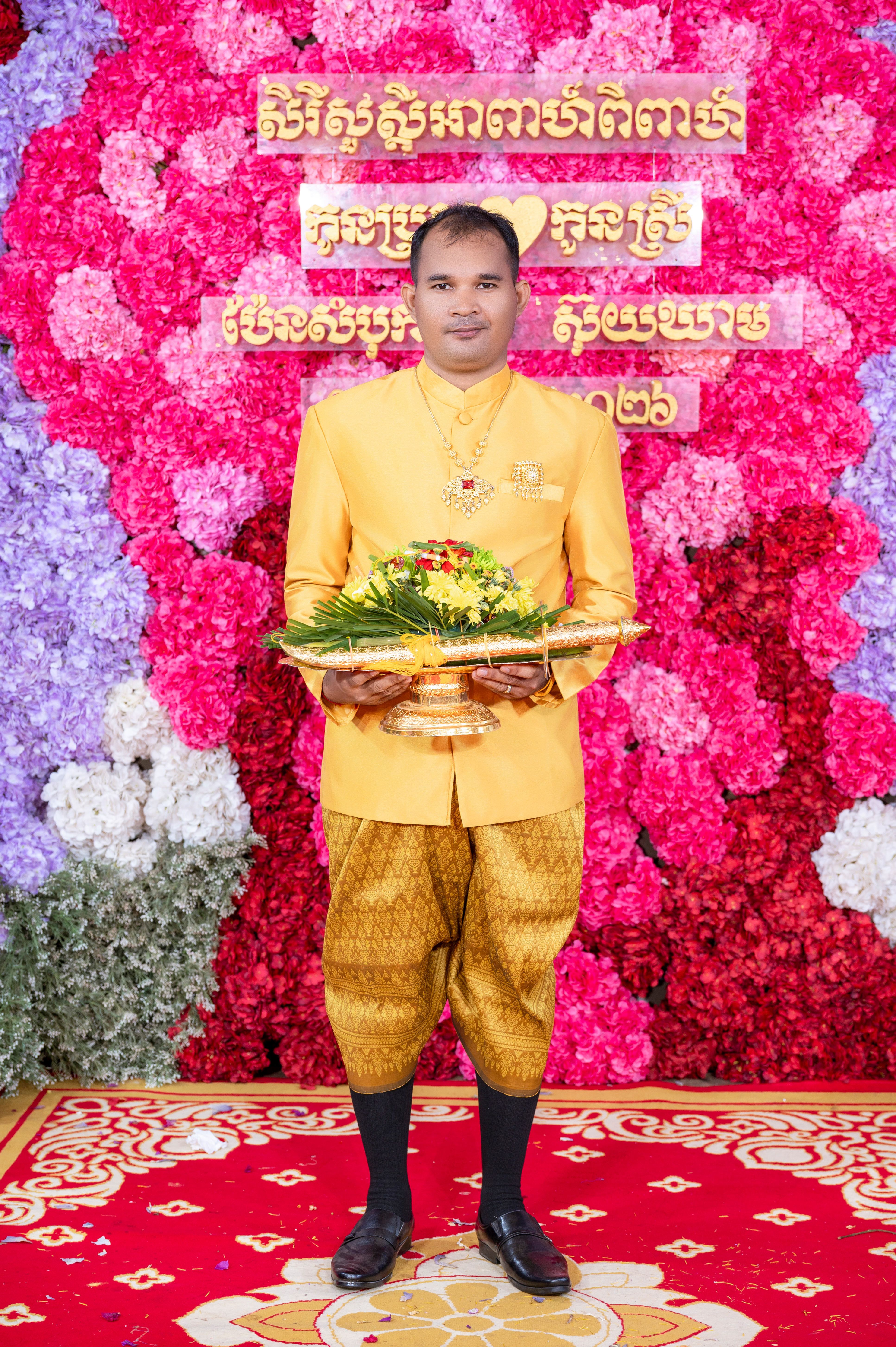 A man in traditional attire poses during a Cambodian wedding ceremony against a vibrant floral backdrop.