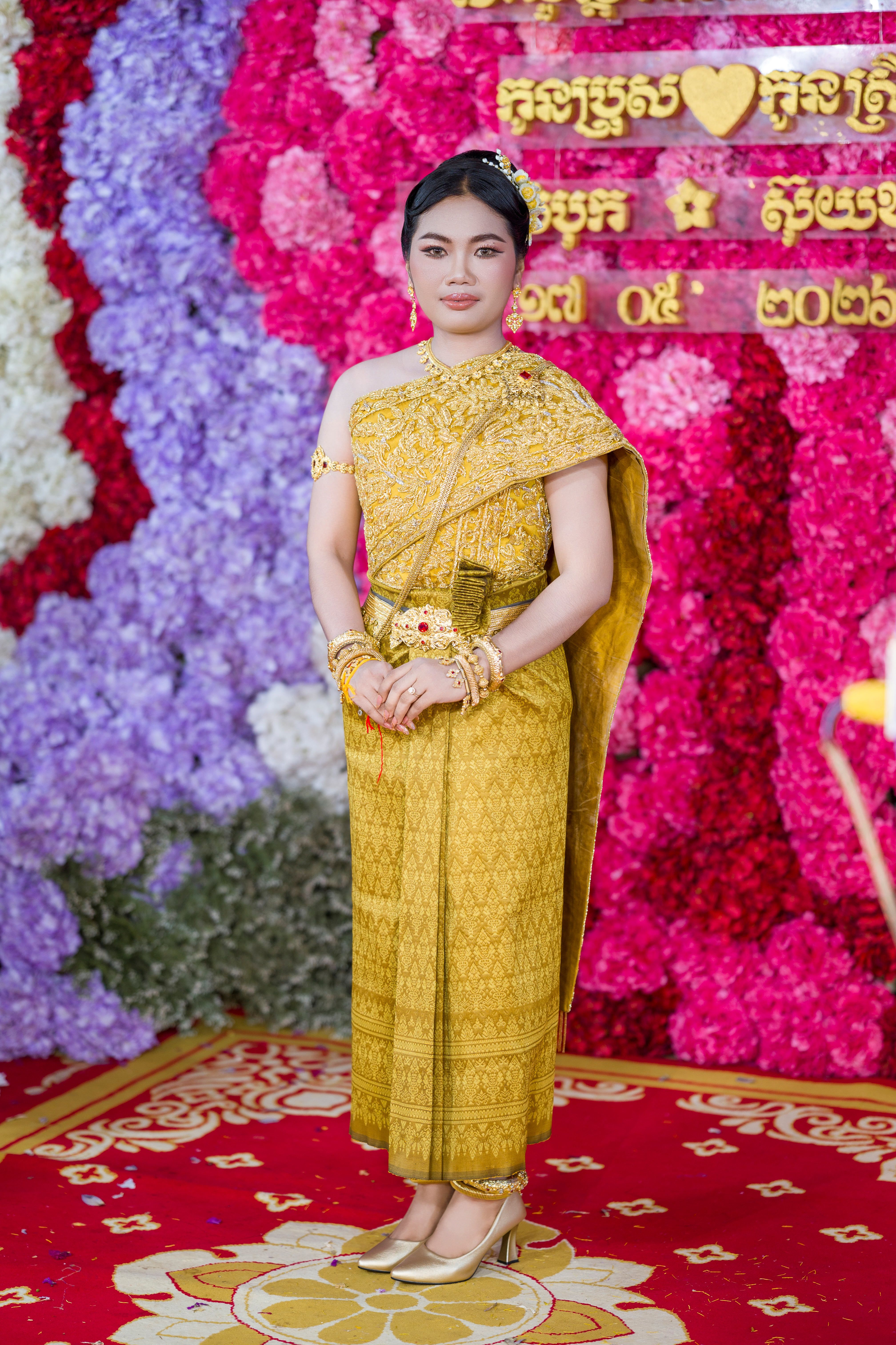 A woman in traditional Cambodian wedding attire stands in front of a colorful floral backdrop.