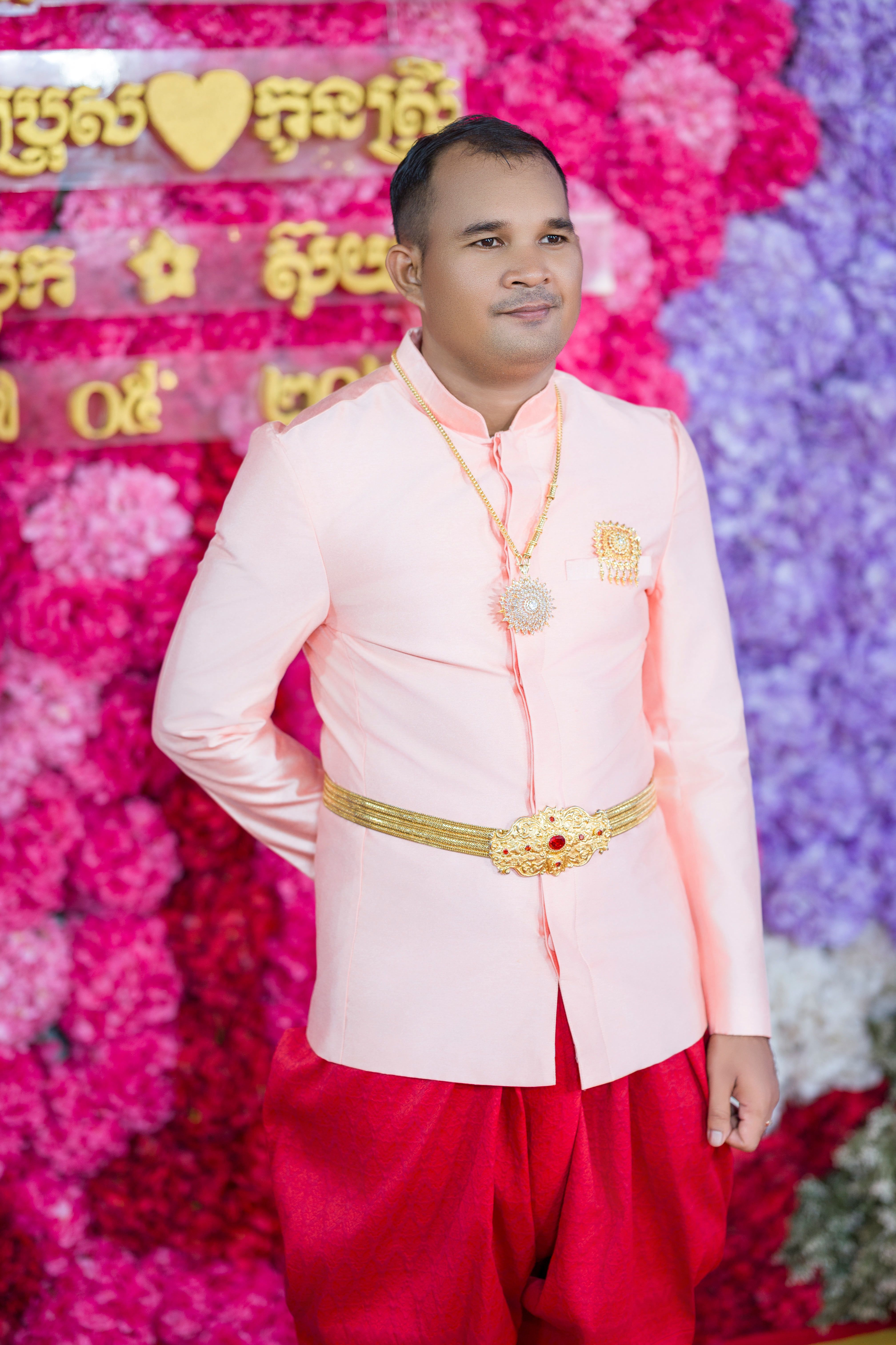 Elegant Cambodian man in traditional wedding attire with a vibrant floral backdrop.