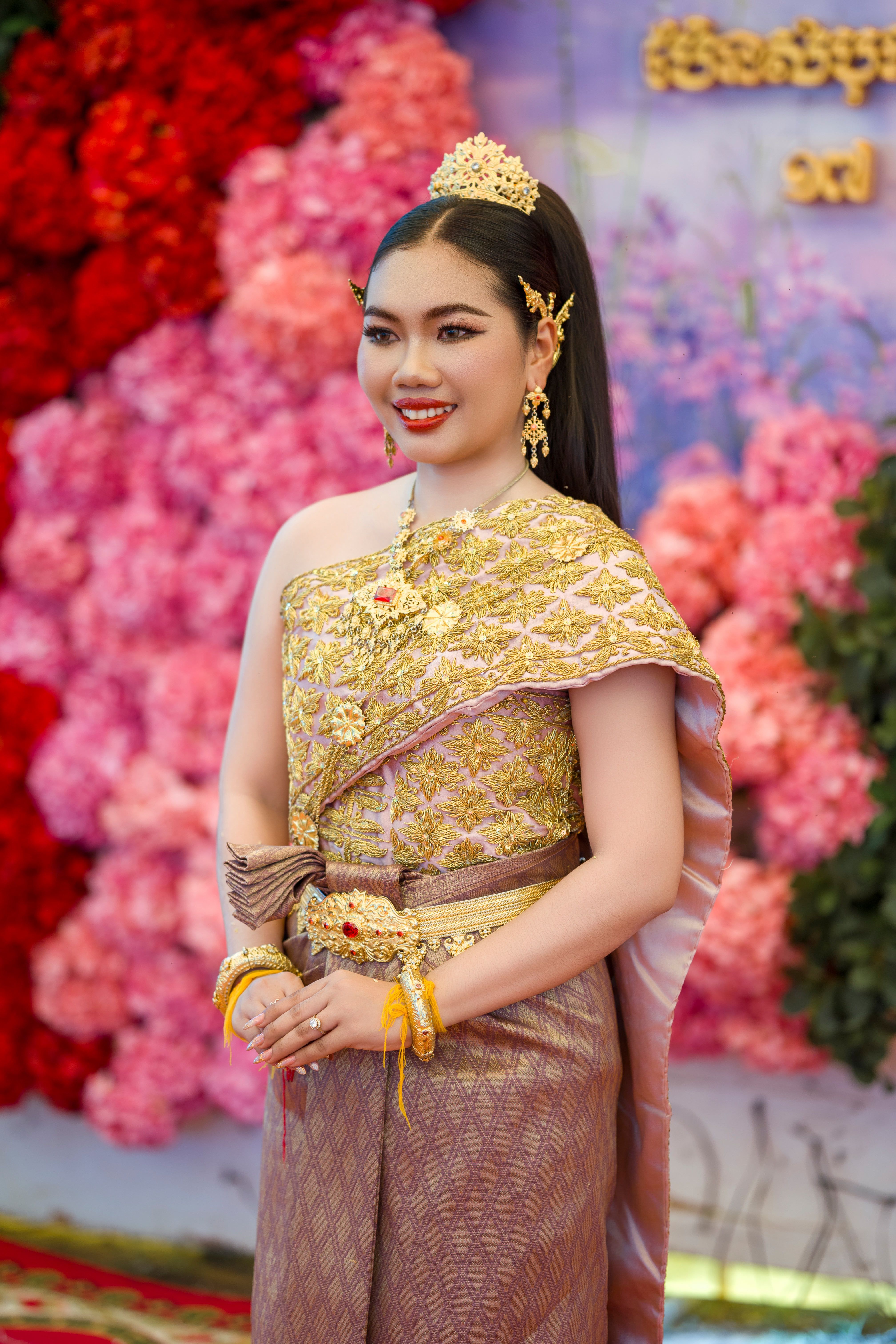 A woman in ornate traditional Cambodian wedding attire stands before a floral backdrop.