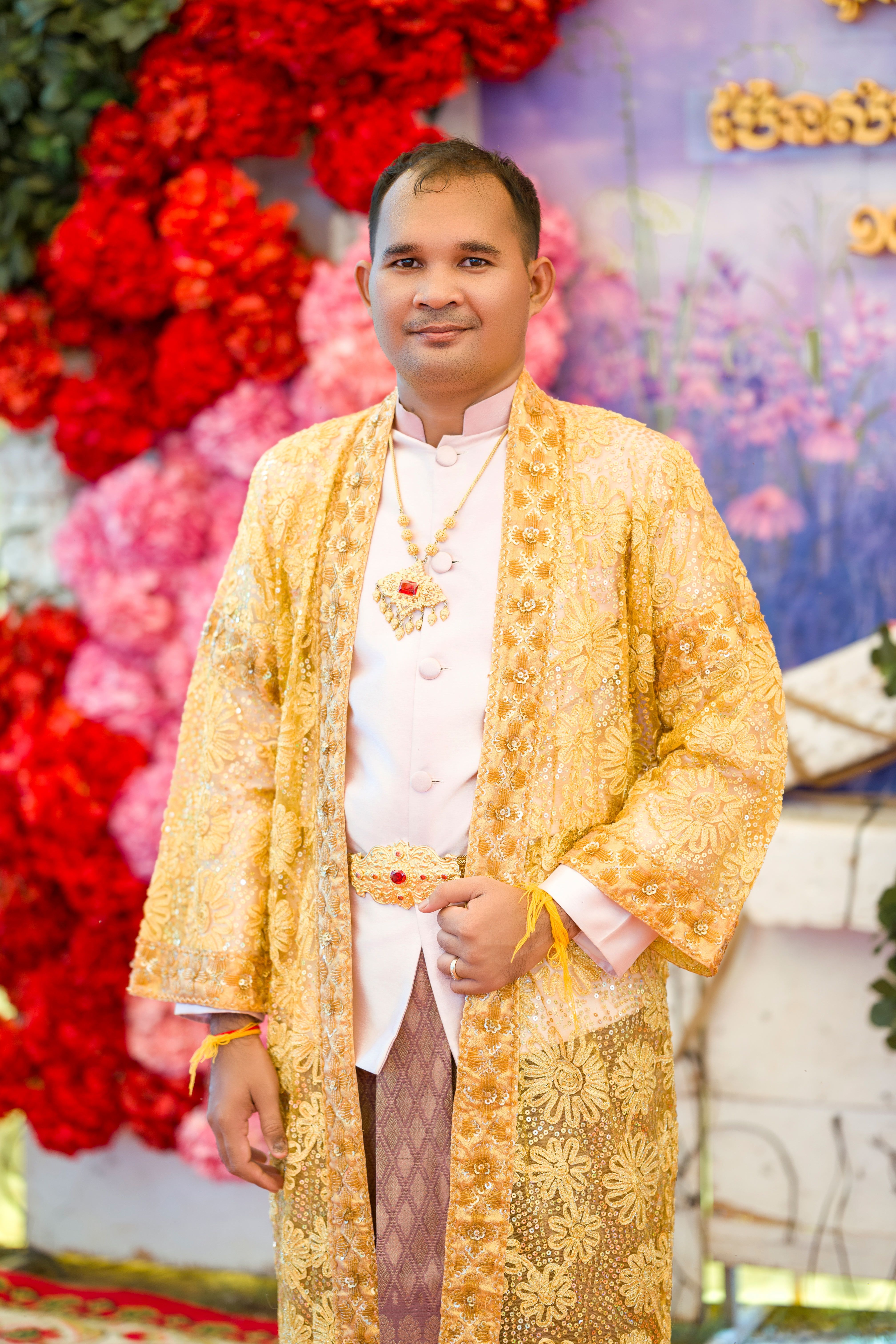 A man in traditional Khmer wedding attire stands in front of a vibrant floral backdrop in Cambodia.