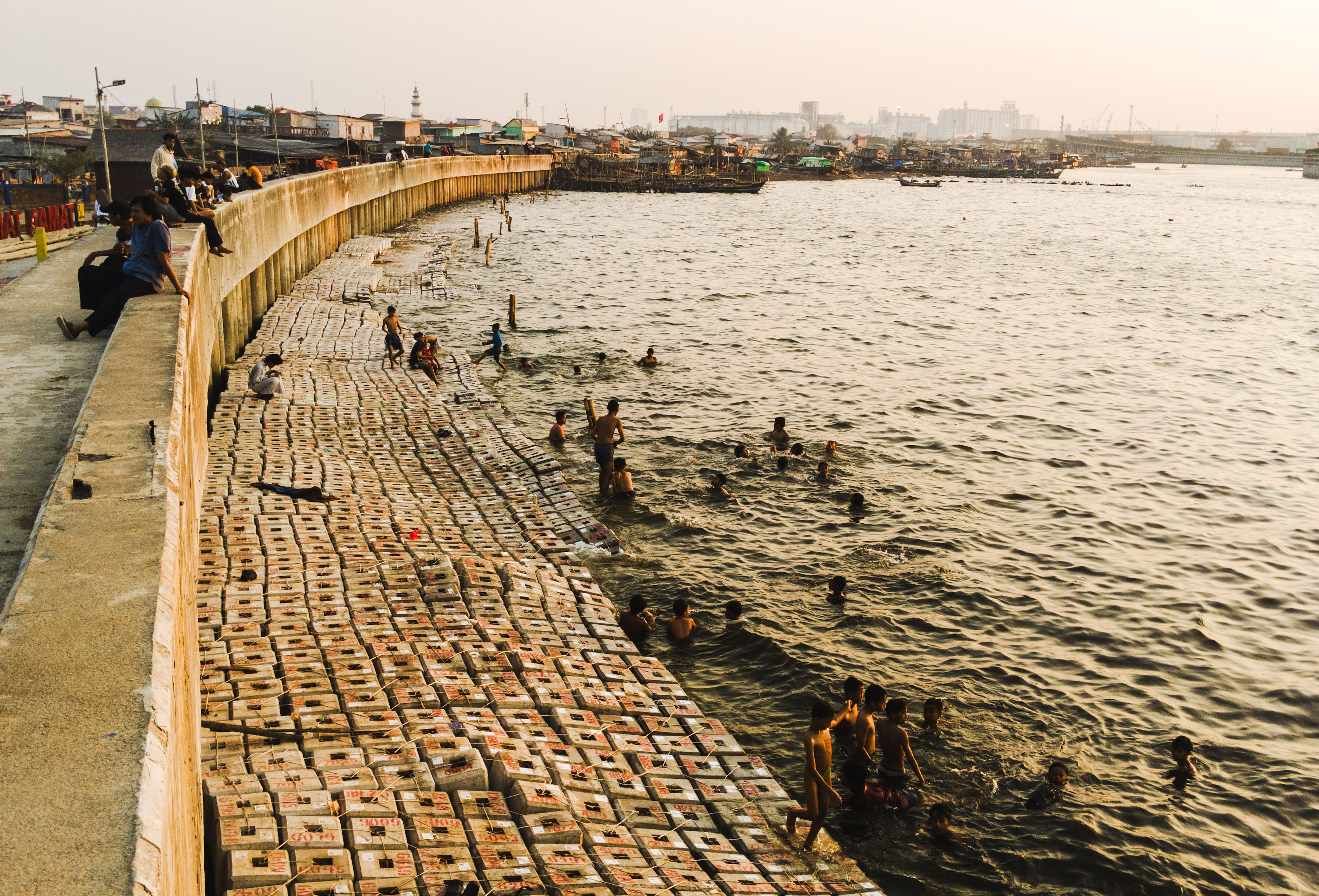 Adults and children swim by a waterfront promenade in North Jakarta, Indonesia during a warm day.