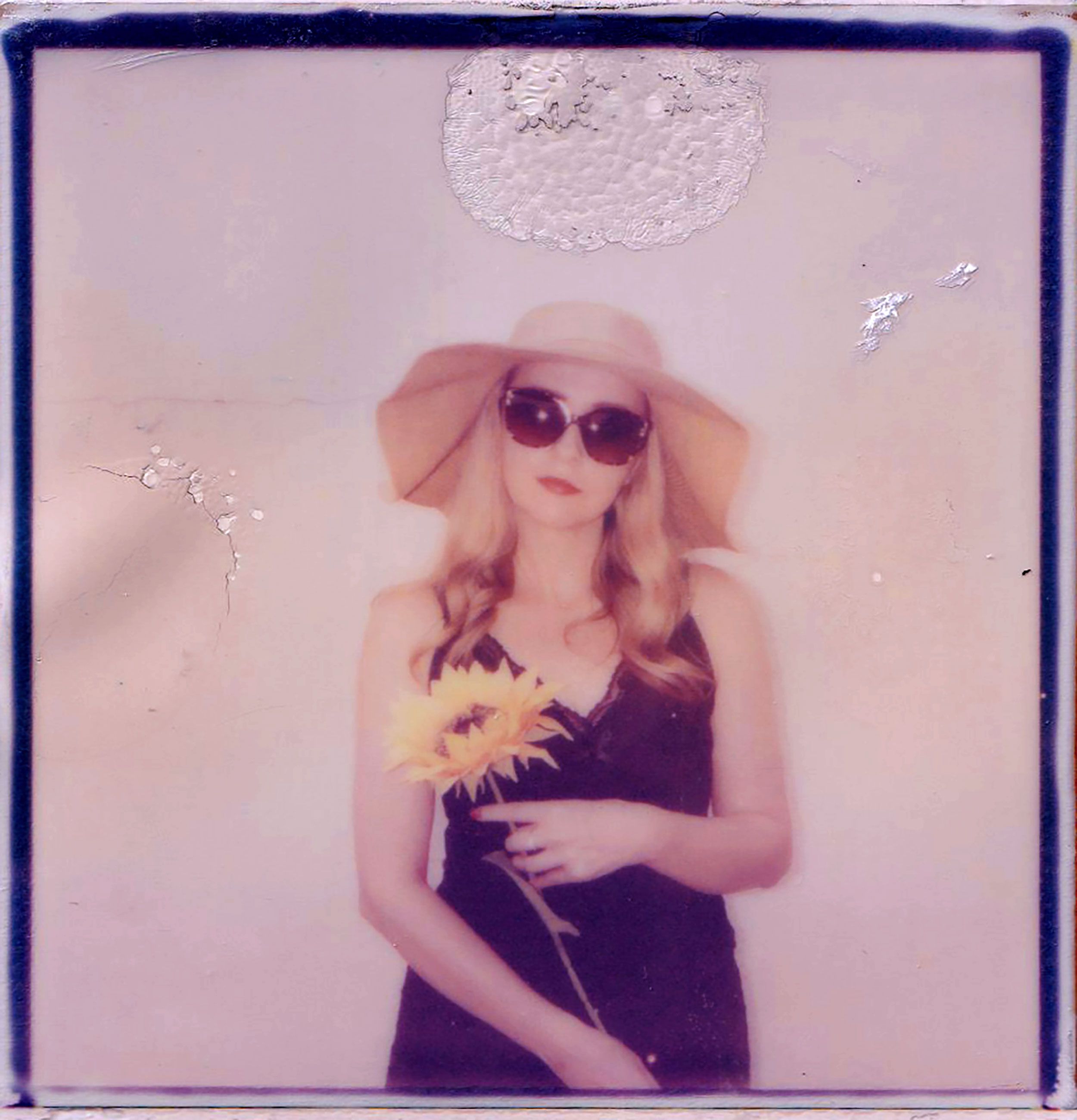 A woman in sunglasses and hat holds a sunflower in a vintage Polaroid photo.
