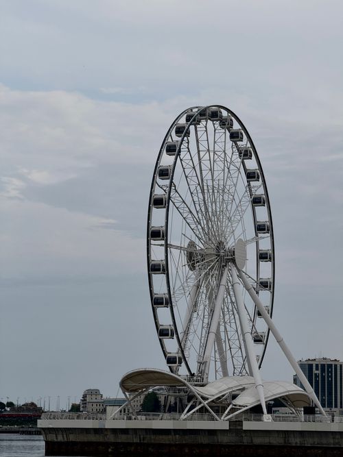 Free Ferris wheel by the Caspian Sea in Baku, showcasing an overcast sky and architectural design. Stock Photo