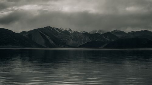 Free A moody and dramatic landscape of mountains reflected in water under a cloudy sky. Stock Photo