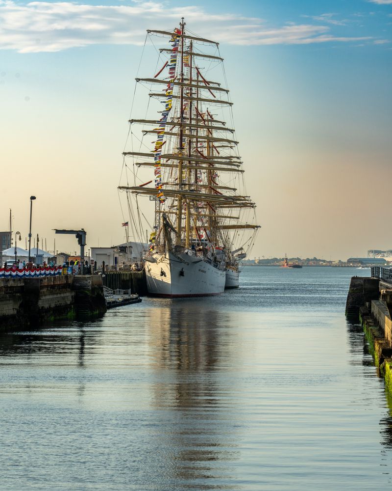 Tall ship silhouetted at golden hour in a harbour, crews gathering at the railing at dusk