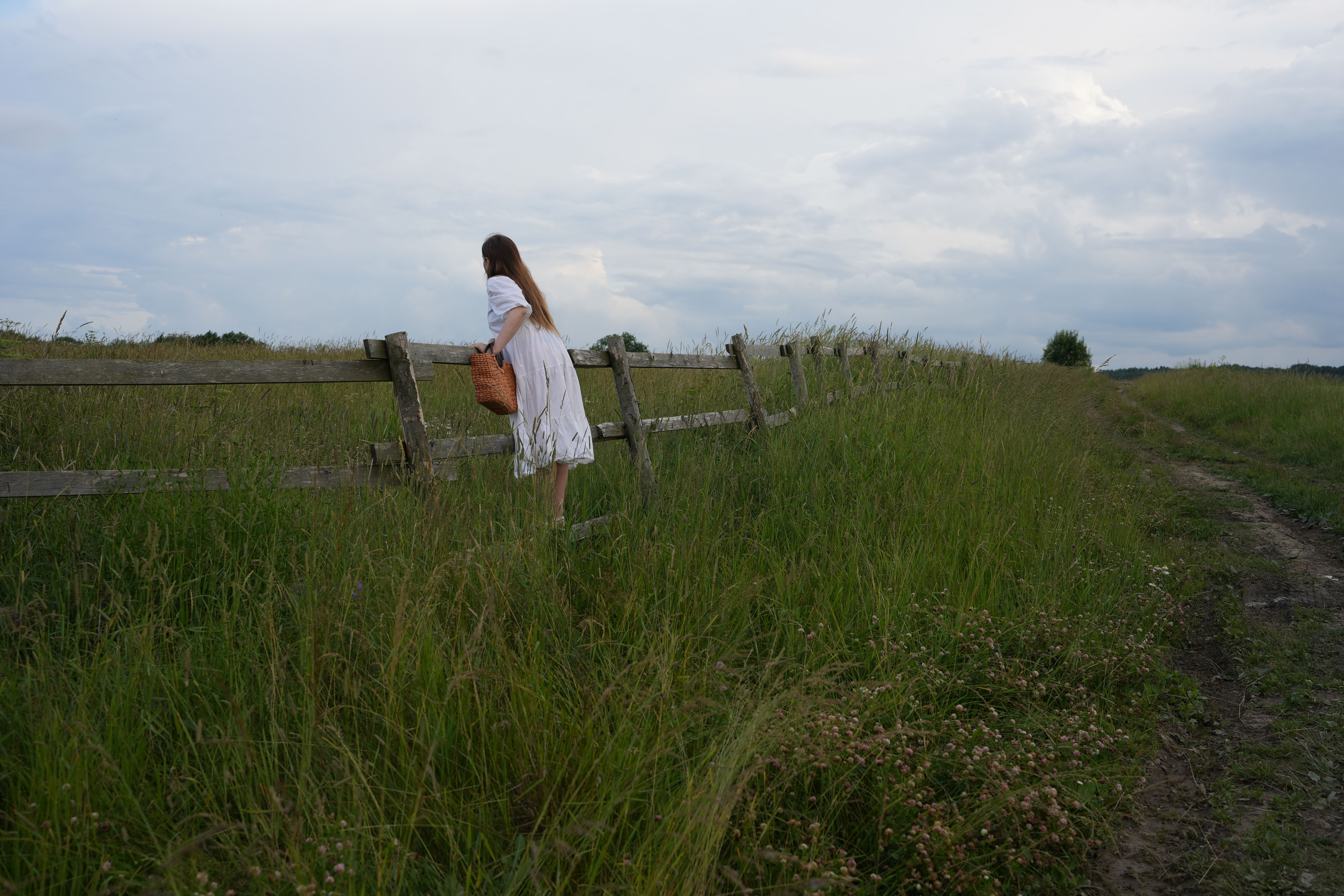 A woman in a white dress walks alongside a wooden fence in a lush, green countryside field under a cloudy sky.