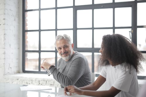 Happy aged gray haired coach in casual wear looking at camera while supporting young African American coworker in office boardroom