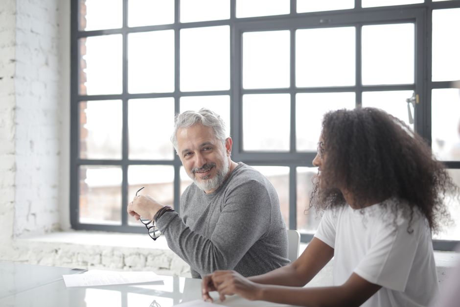 Happy aged gray haired coach in casual wear looking at camera while supporting young African American coworker in office boardroom