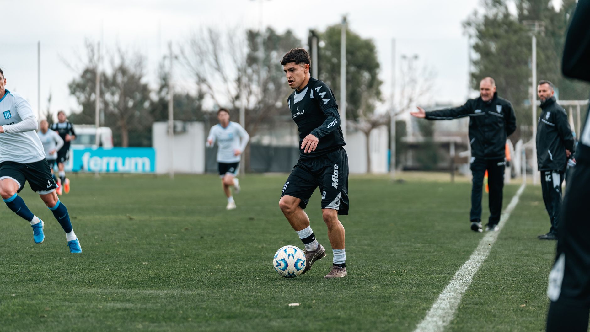 A football coach timing a dribbling drill beside cones, players resting near a tactical board