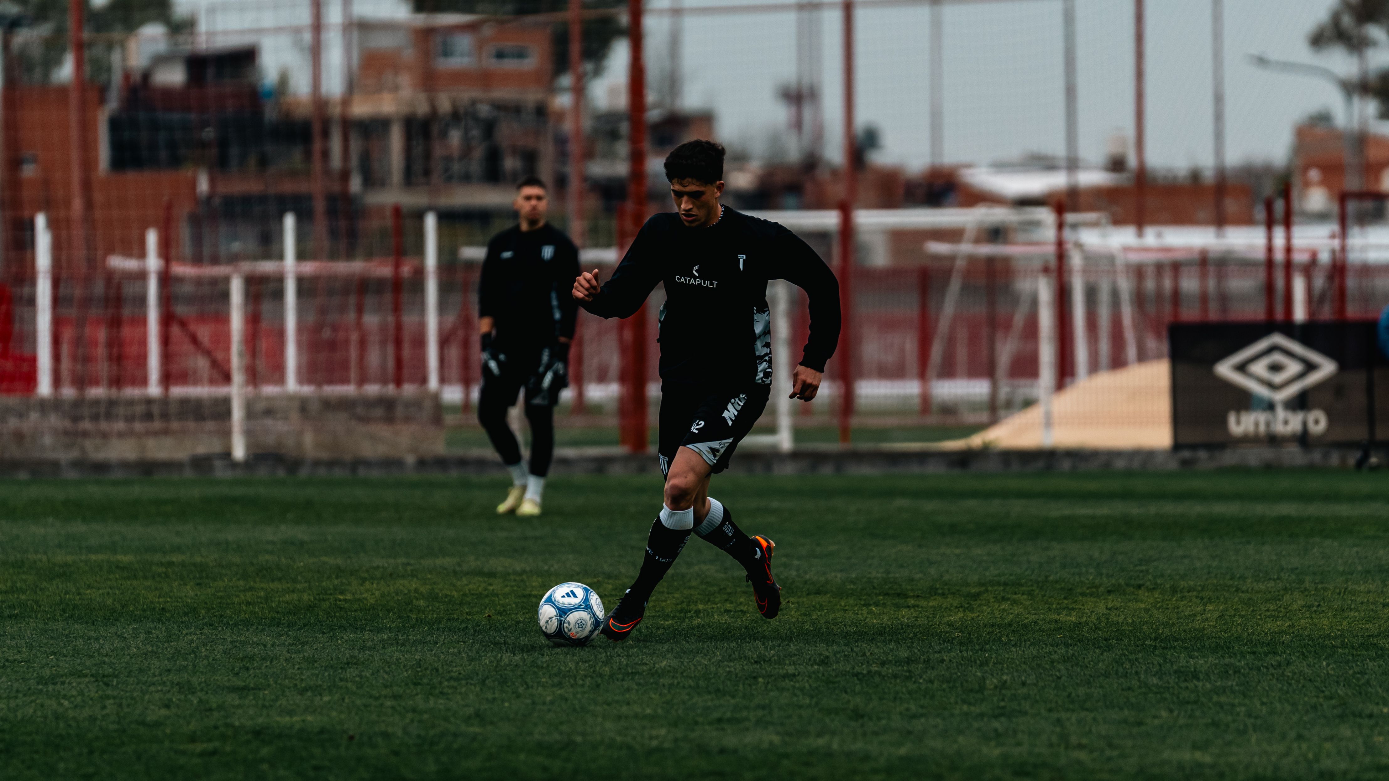 Güneşli bir havada antrenman sahasında top süren bir futbolcu.