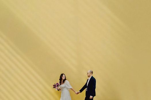 A happy couple holding hands against a sunlit yellow wall, capturing love and harmony.