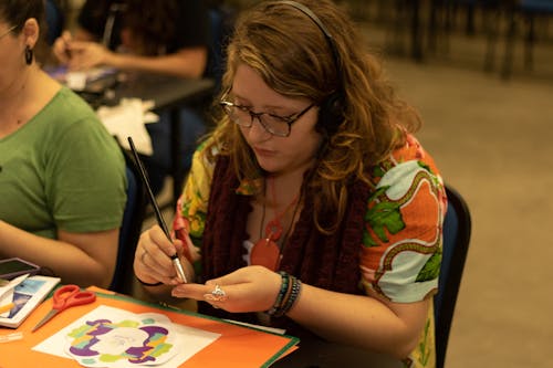 High angle young attentive female artist in casual clothes and headphones gluing paper using paintbrush while creating picture during lesson