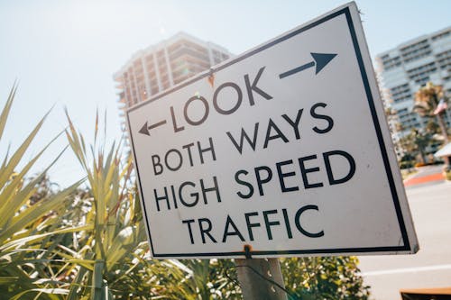 Direction sign with arrow and look both ways high speed traffic text placed on sidewalk near tropical plants against modern city buildings