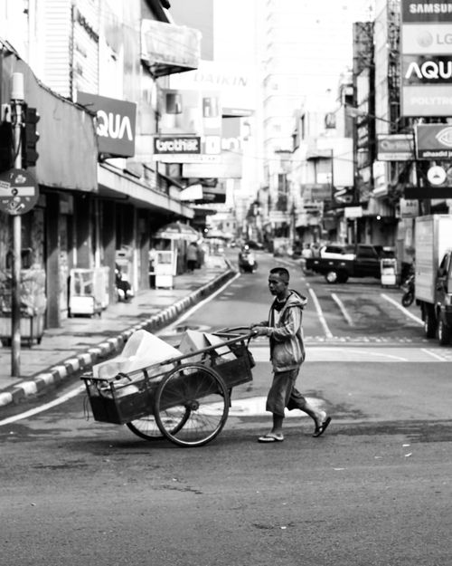 Free A black and white street scene capturing daily life in Bandung, Indonesia with a man pushing a cart. Stock Photo