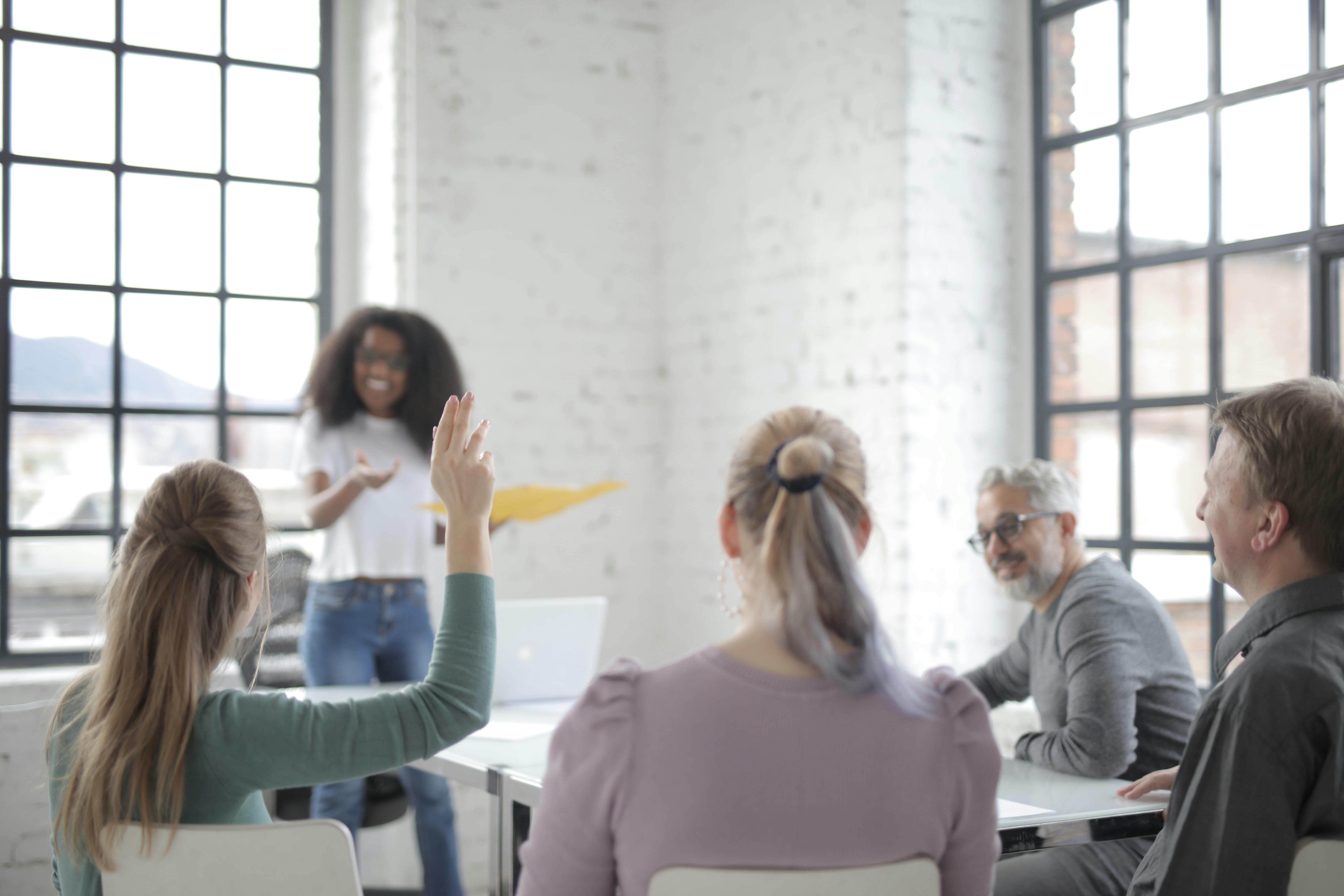 Female worker in casual wear raising hand for asking question during corporate diverse group meeting in modern office boardroom