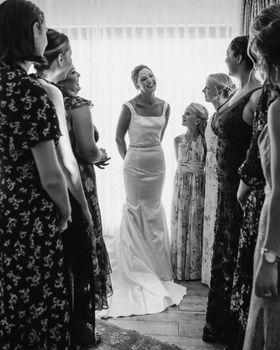 A smiling bride in a wedding dress surrounded by bridesmaids in an intimate indoor setting.