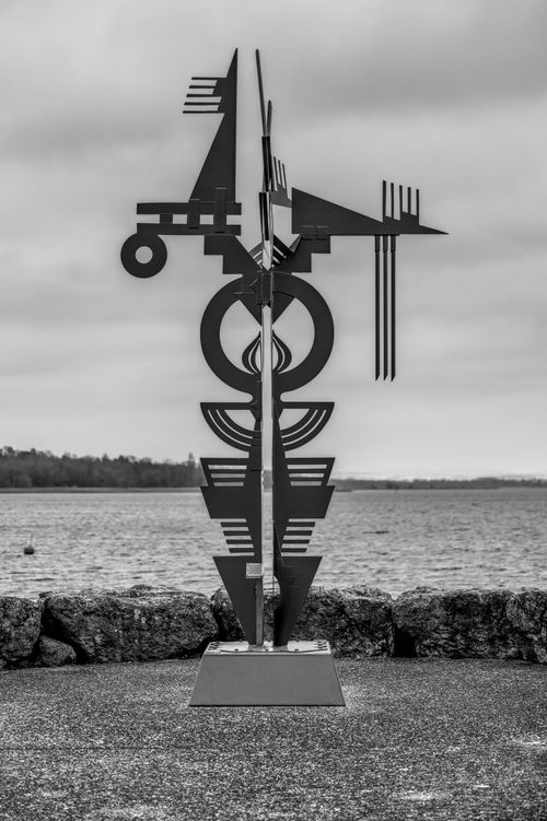 Free Intricate abstract metal sculpture displayed near a tranquil lake under a cloudy sky. Stock Photo
