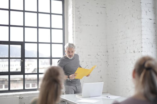 Serious aged gray haired bearded male speaker in casual wear answering question at meeting in modern office boardroom while standing against table with laptop and presenting ideas to colleagues