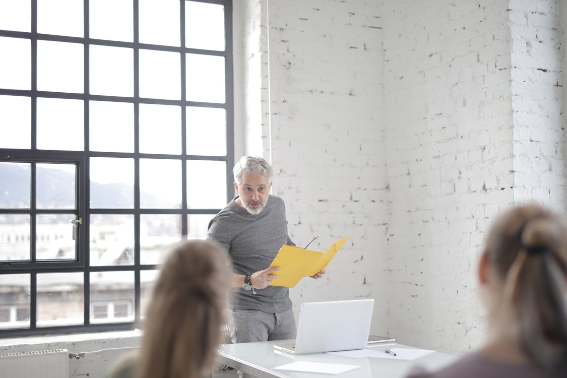 Free Serious aged gray haired bearded male speaker in casual wear answering question at meeting in modern office boardroom while standing against table with laptop and presenting ideas to colleagues Stock Photo