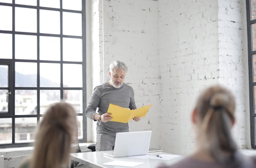 Focused aged gray haired bearded male speaker in casual wear presenting ideas to colleagues in conference room at meeting while standing at table with laptop