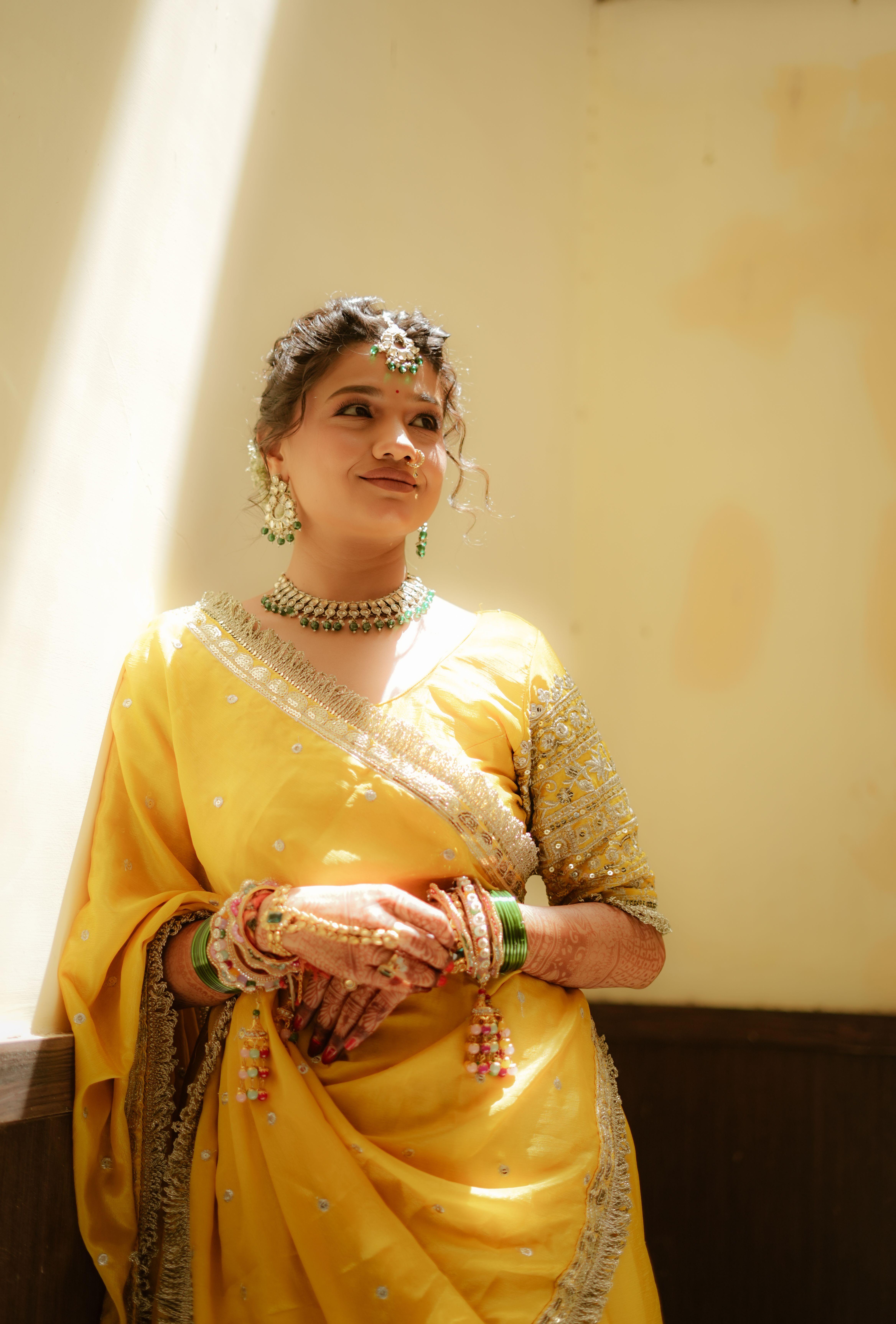 Portrait of a woman in a yellow saree with traditional jewelry. Sunlit indoor setting.