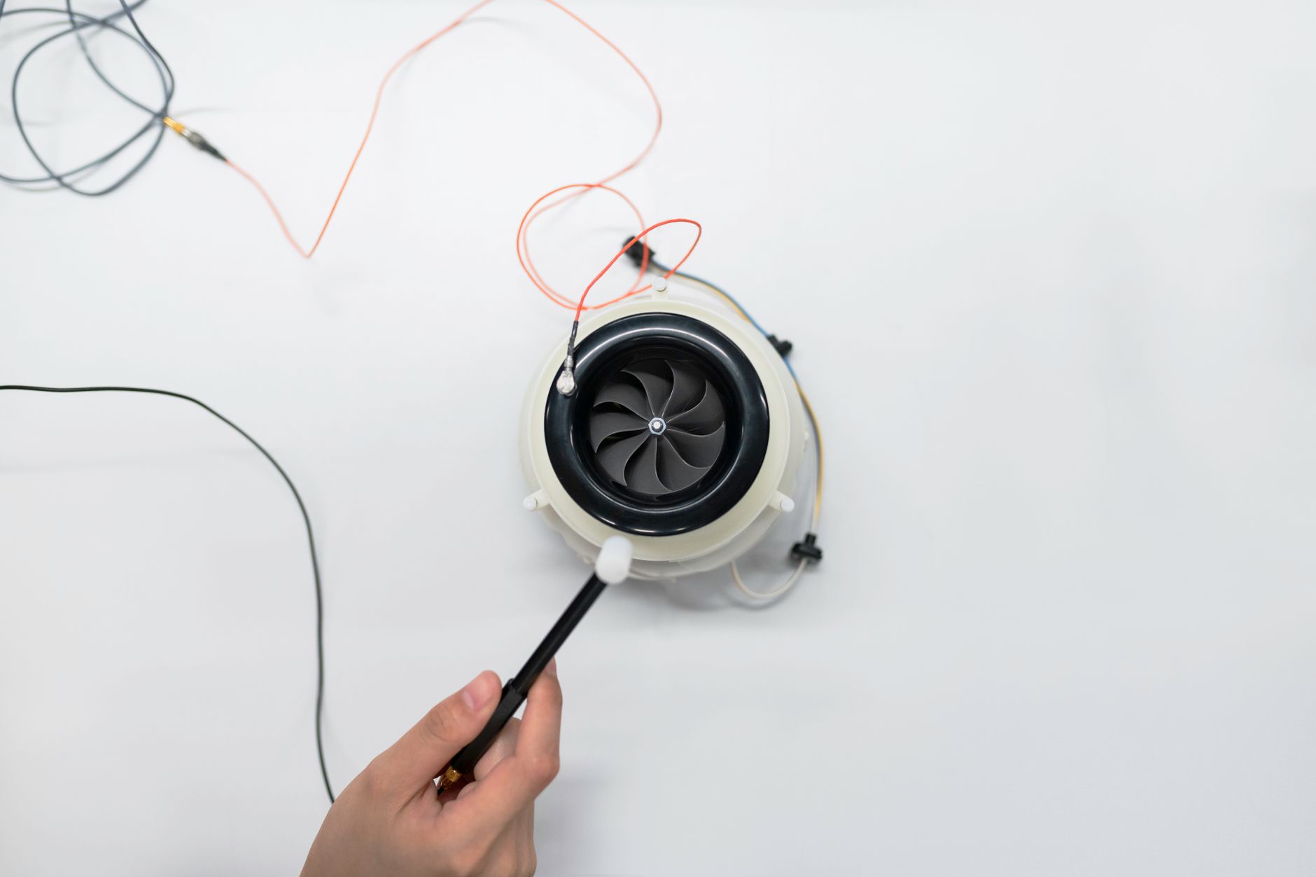 Overhead view of a hand adjusting a small electric fan with wires on a white surface.