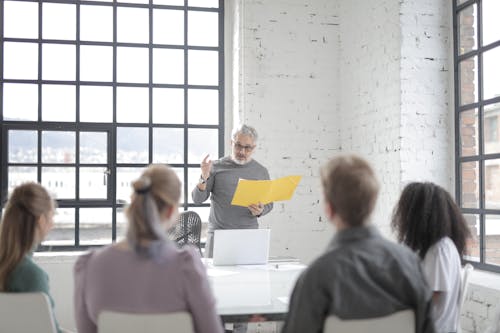 Free Serious middle aged executive in eyeglasses talking to diverse subordinates while taking yellow folder and standing in front of open laptop in light office Stock Photo