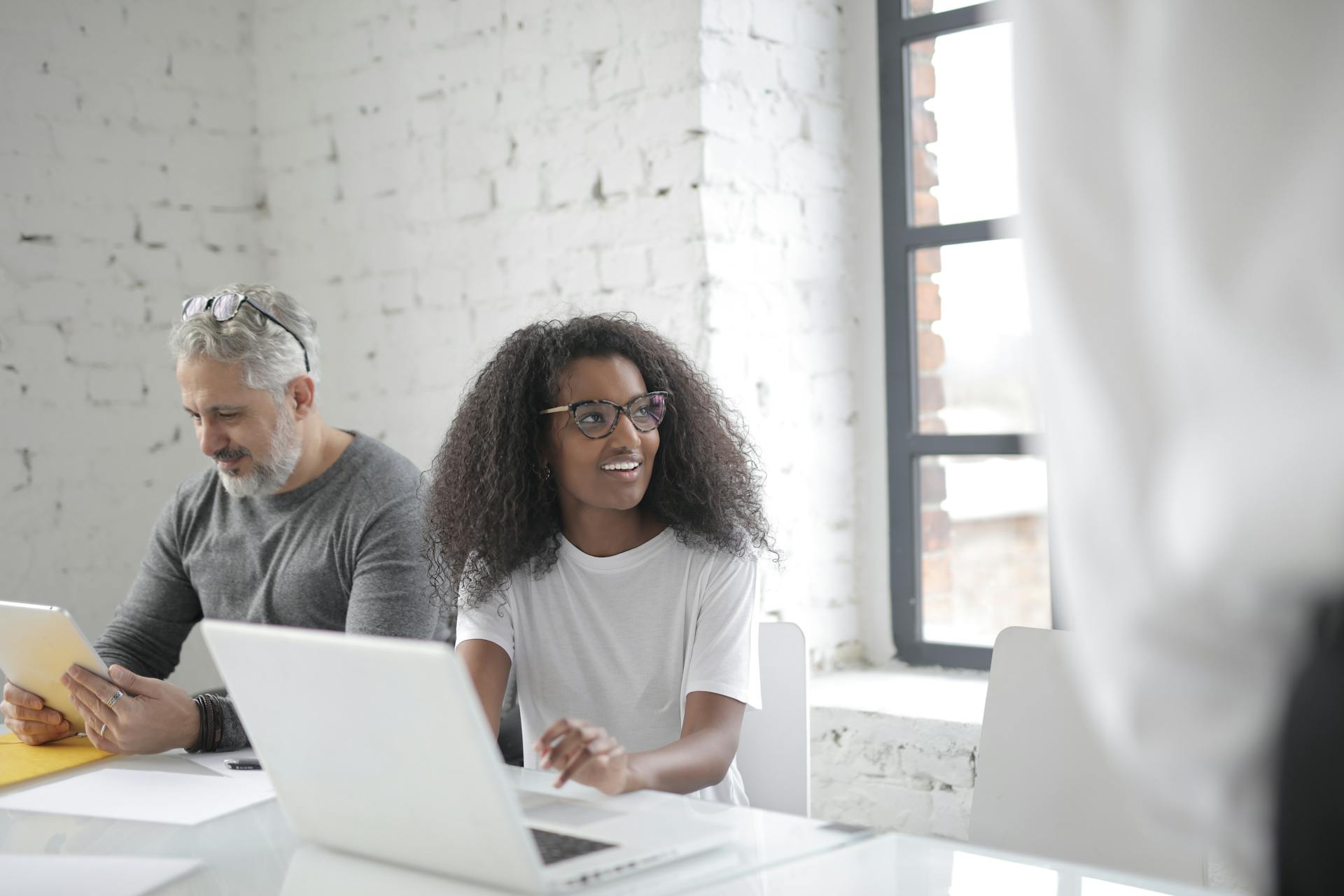 African American female employee working on netbook and looking away while sitting near middle aged coworker watching tablet in light workplace