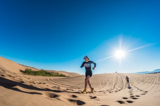 A runner in vibrant attire dashes across the sunlit desert dunes under a clear blue sky.