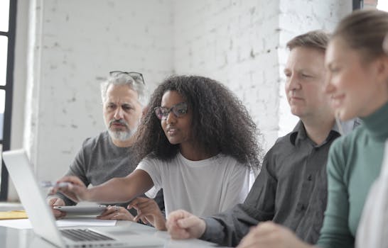 African American female worker in eyeglasses pointing with pen at netbook screen while talking about project to multiethnic partners sitting at table in office in afternoon