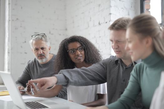 Male employee pointing at portable computer screen while expressing opinion about work project to diverse colleagues sitting at desk in office