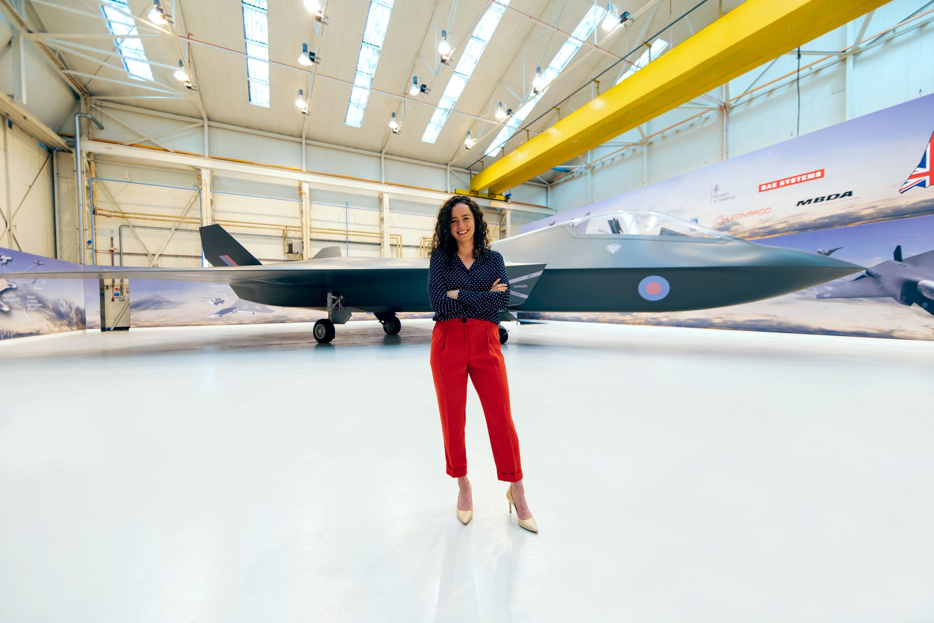 A female engineer stands confidently in front of a modern fighter jet in a hangar.