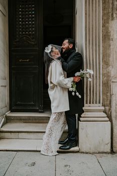 Bride and groom share a romantic moment on a vintage doorstep, holding flowers.