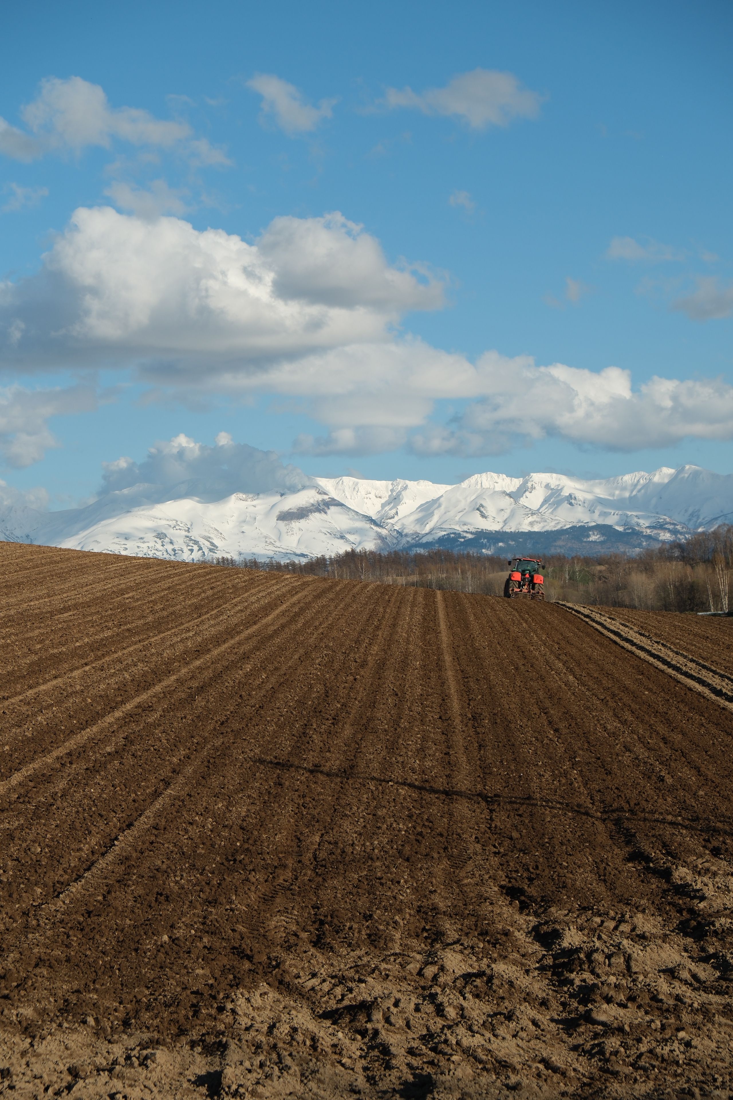 A tractor plows a vast field under a vivid blue sky with snowy mountains in Hokkaido.