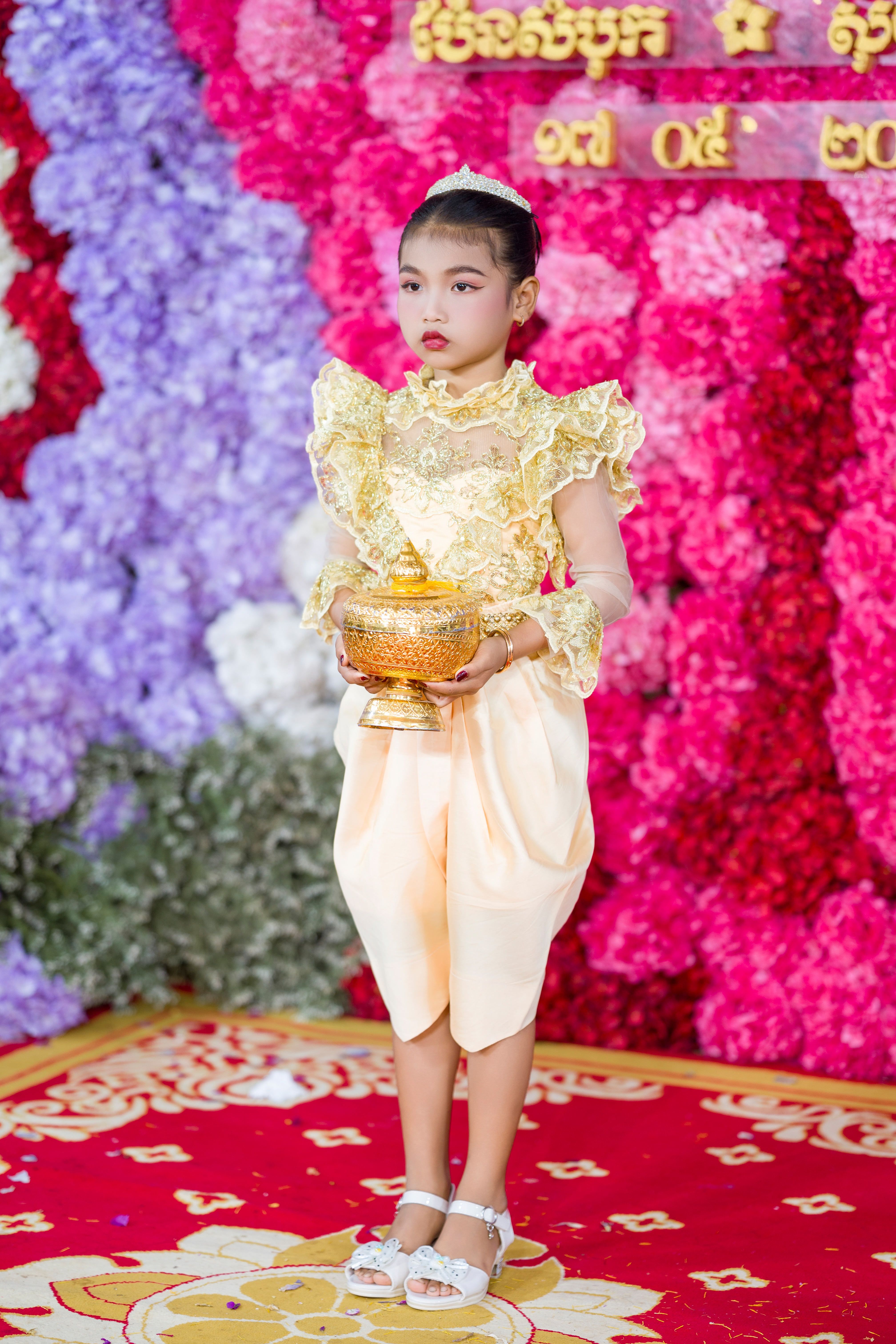 A child in ornate Cambodian dress holding a golden bowl, with a floral backdrop.