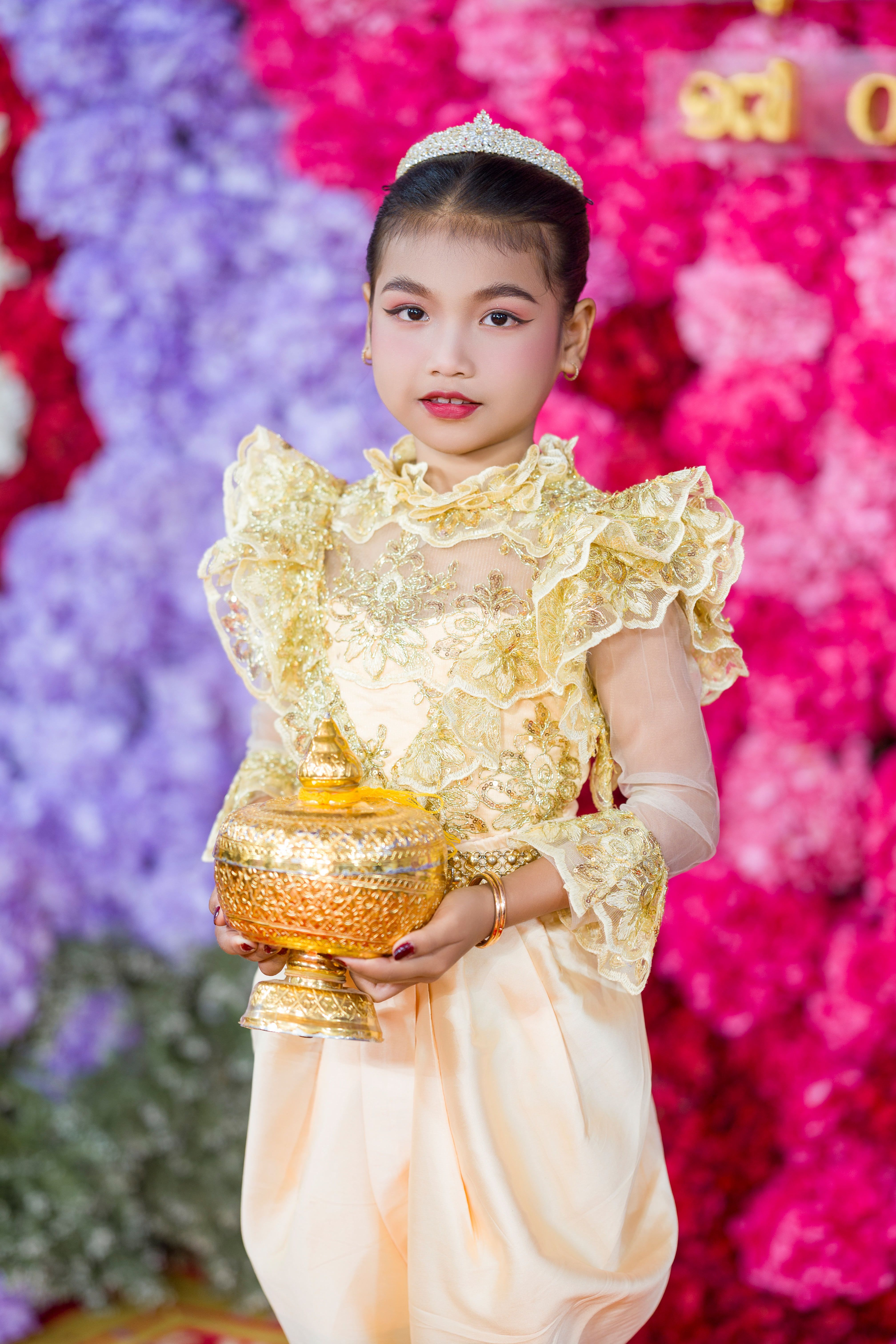 Girl in Cambodian traditional attire holding a golden bowl with a vibrant floral background.