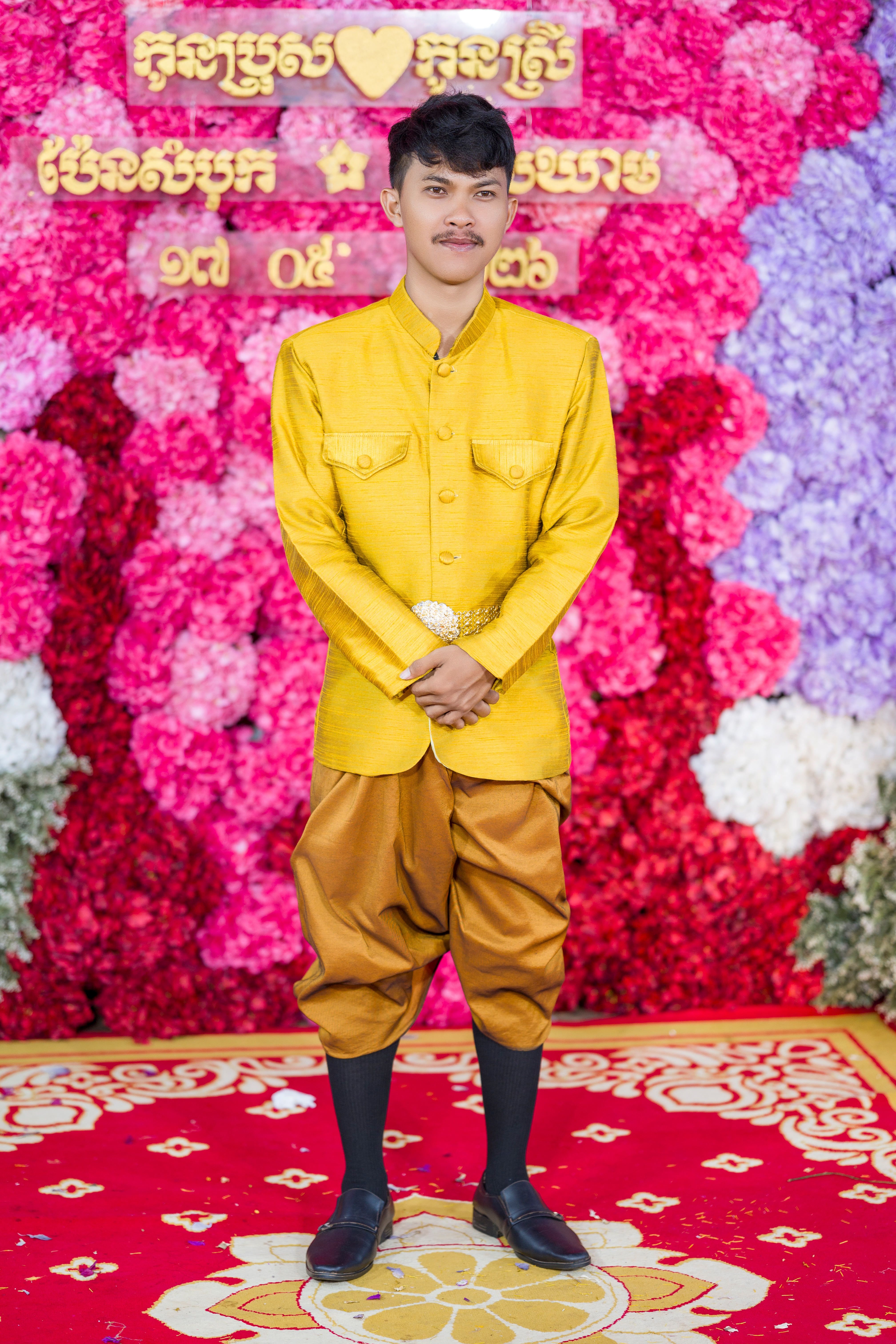 A young man in traditional Khmer wedding attire stands against a floral backdrop in Cambodia.