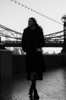 Black and white photo of a woman walking near Tower Bridge in London at twilight.