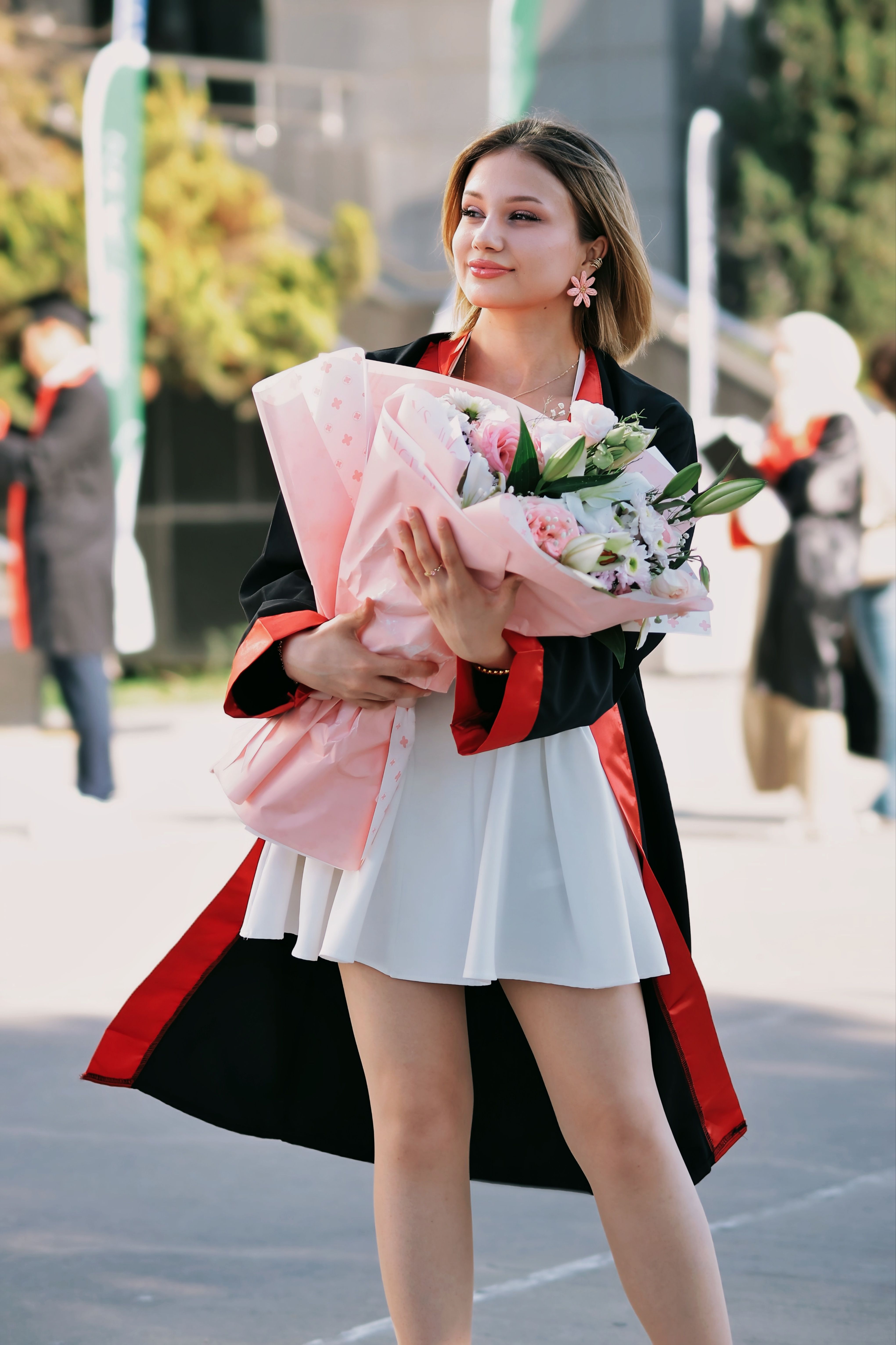 A young woman celebrates her graduation outdoors, holding a bouquet of flowers.