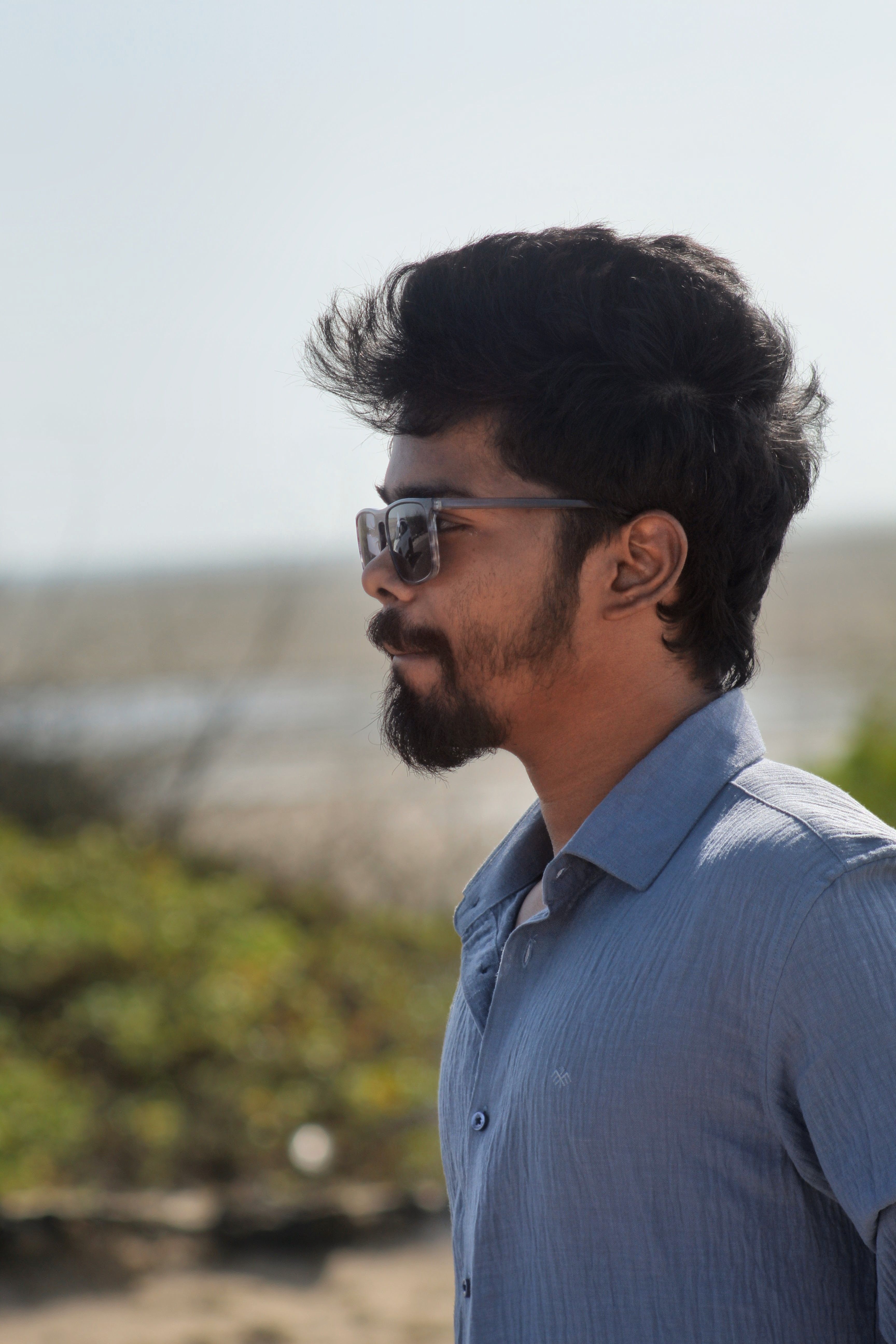Side profile of a man wearing sunglasses, standing outdoors at the beach on a sunny day.