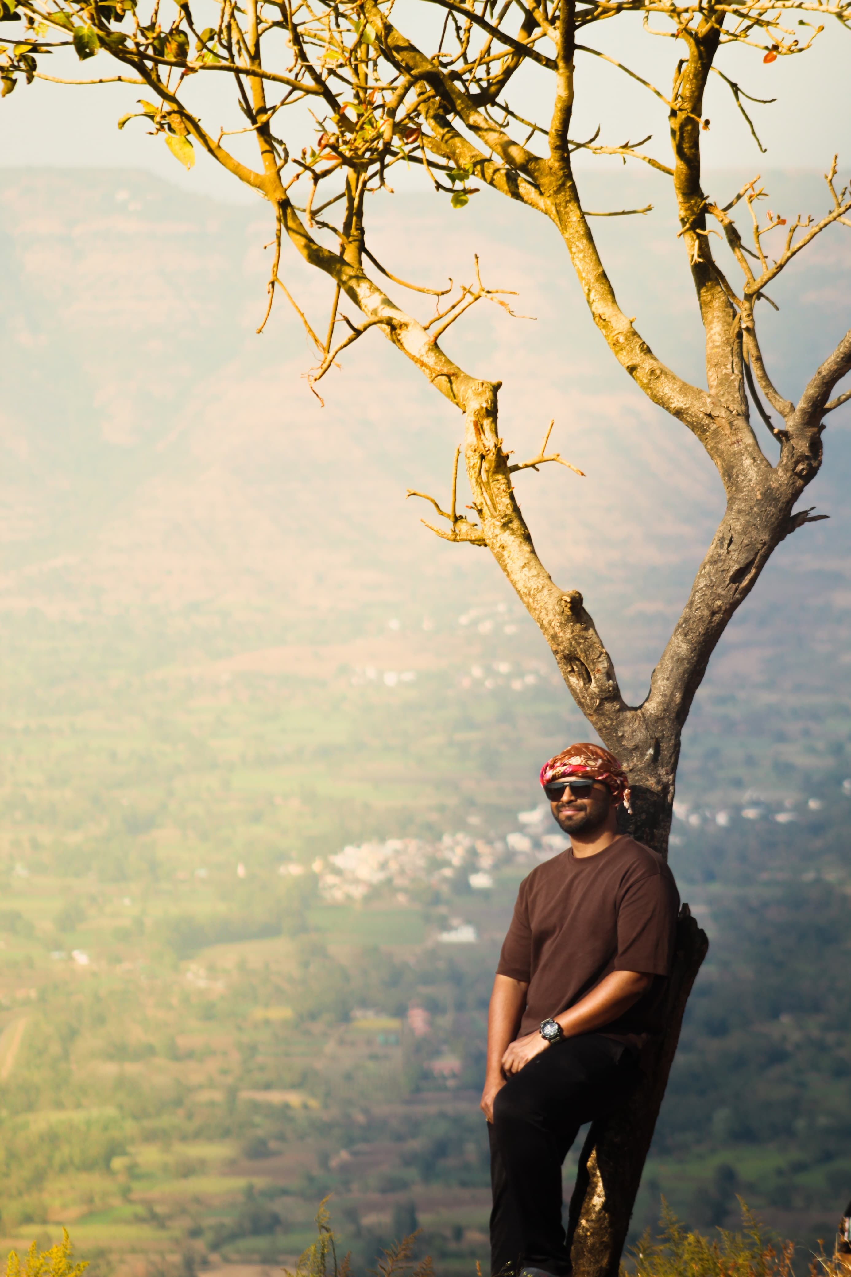 Man in sunglasses and headband relaxes by a tree overlooking a scenic valley.