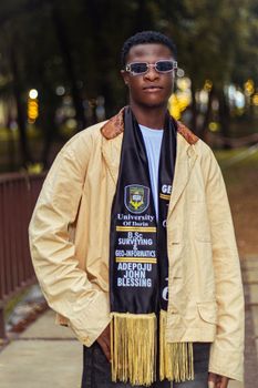 A stylish graduate from University of Ilorin poses confidently outdoors wearing academic scarf and sunglasses.