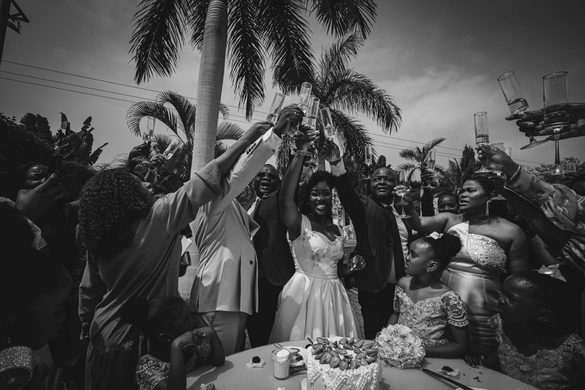 Black and white photo of a lively wedding celebration outdoors in Luanda, Angola with people raising glasses.