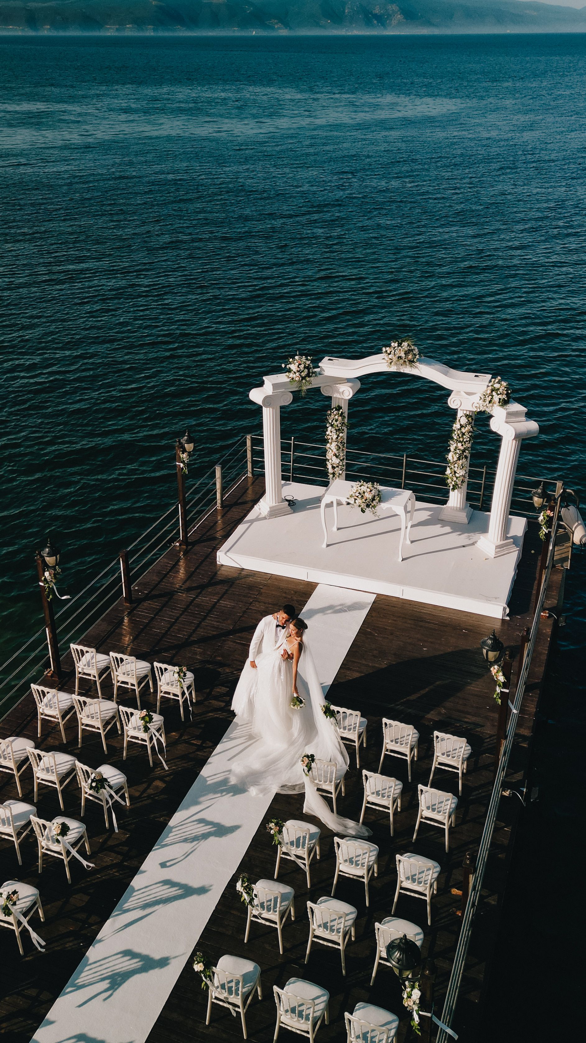 Elegant outdoor wedding setup on a pier by the water, featuring floral decor and an arch.