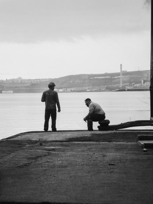 Free Black and white image of two workers by a waterfront in an industrial area. Stock Photo