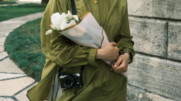 An individual in a green coat holds white flowers and a vintage camera on a walkway.